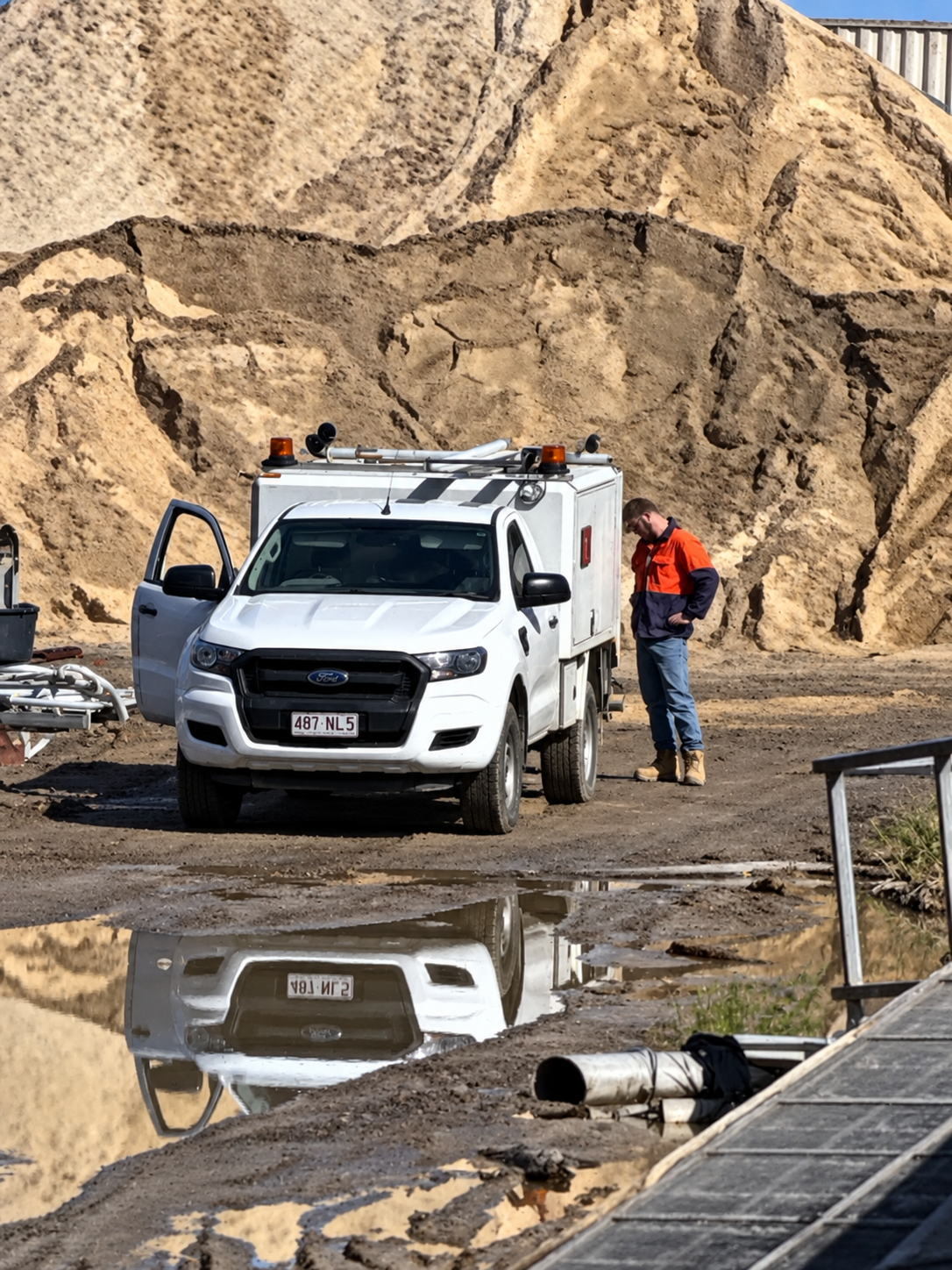 Fabworkz tradesperson beside a white service ute at an industrial site, reflected in a puddle